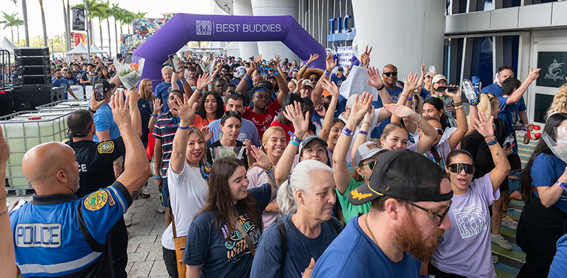 Thousands put their best foot forward at Ed Ansin Best Buddies Friendship Walk at loanDepot Park - Top image Large crowd of participants walking under a purple Best Buddies arch at an outdoor event, with many wearing event t-shirts.